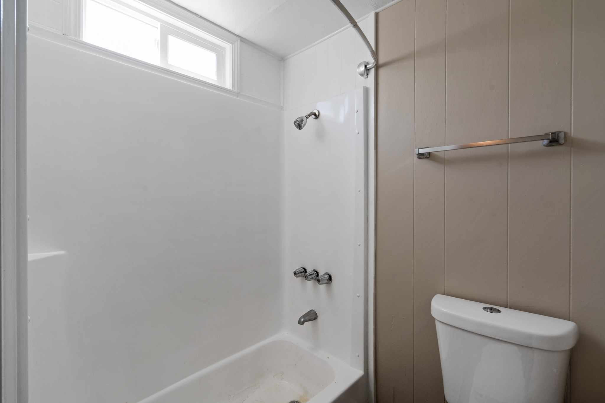 Bathroom with a white bathtub, overhead shower, and a small window above, alongside a toilet and a towel bar on a beige wall.