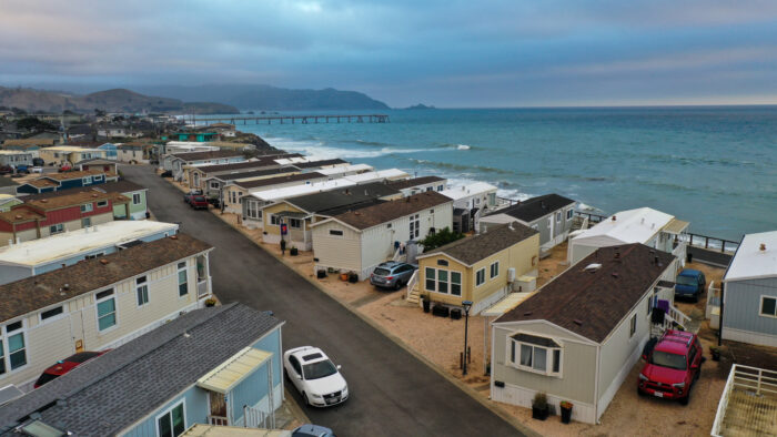 Aerial view of beachfront mobile homes lined along a coastal road with ocean waves and a distant pier in Pacifica, CA