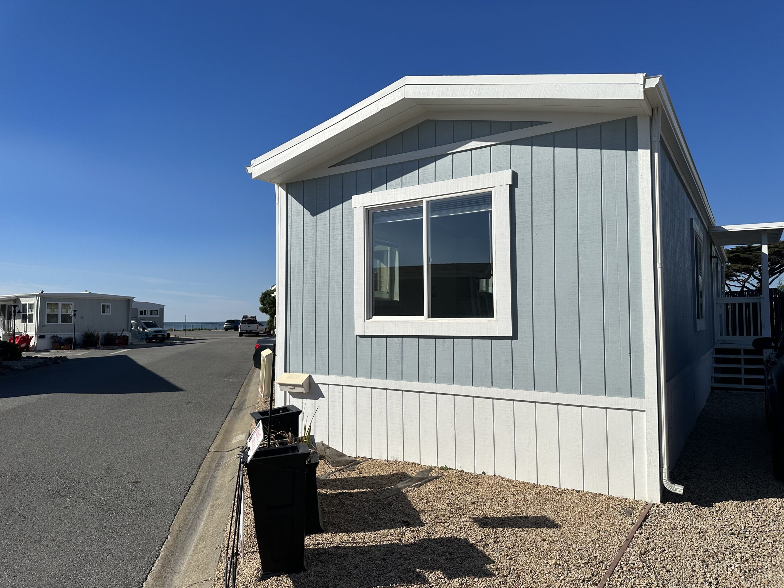 Light blue mobile home with white trim and clear skies, situated in a residential area with a street view.