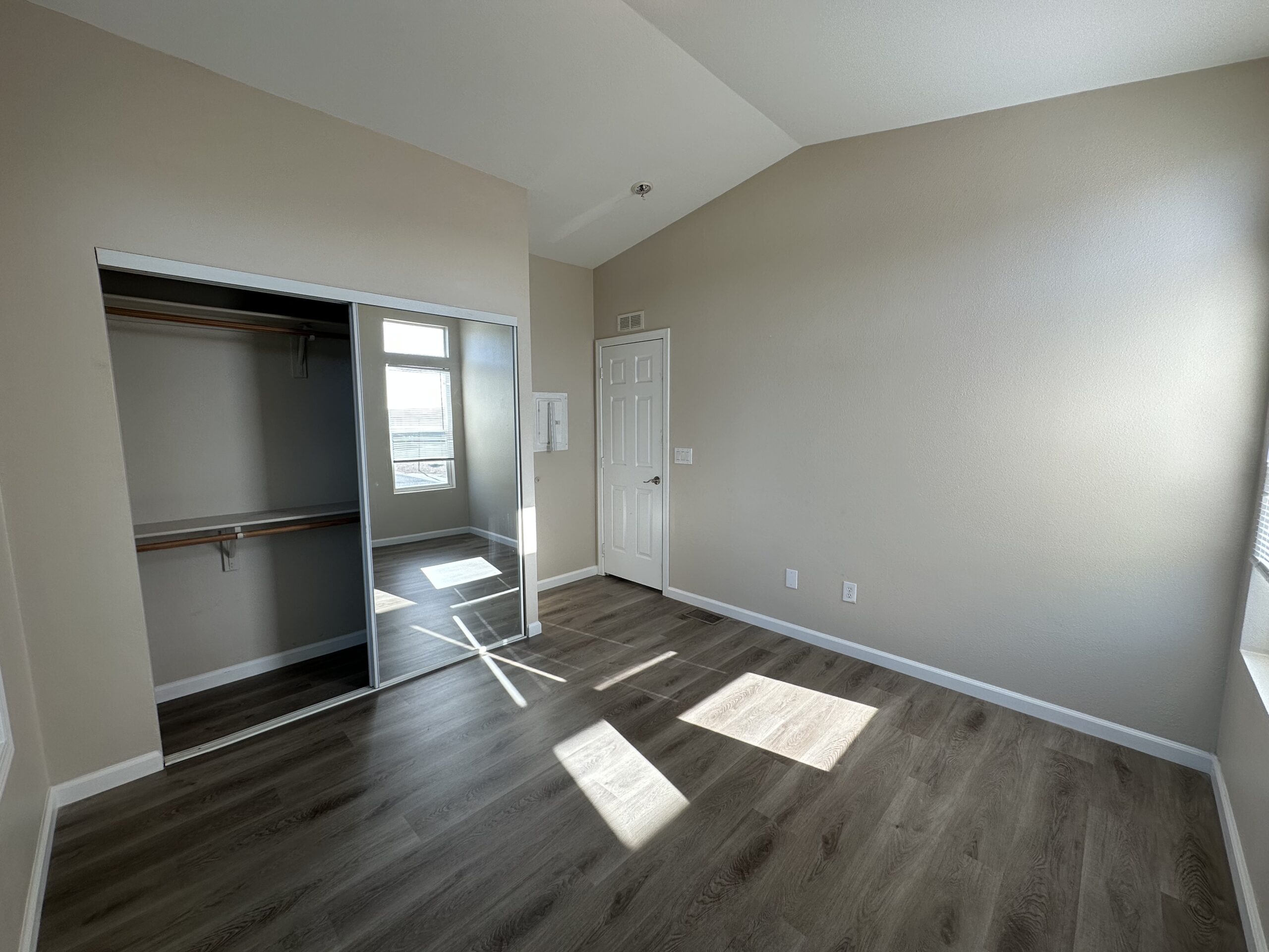 Empty room with an open closet, wood laminate flooring, and a single window casting sunlight on the walls and floor.