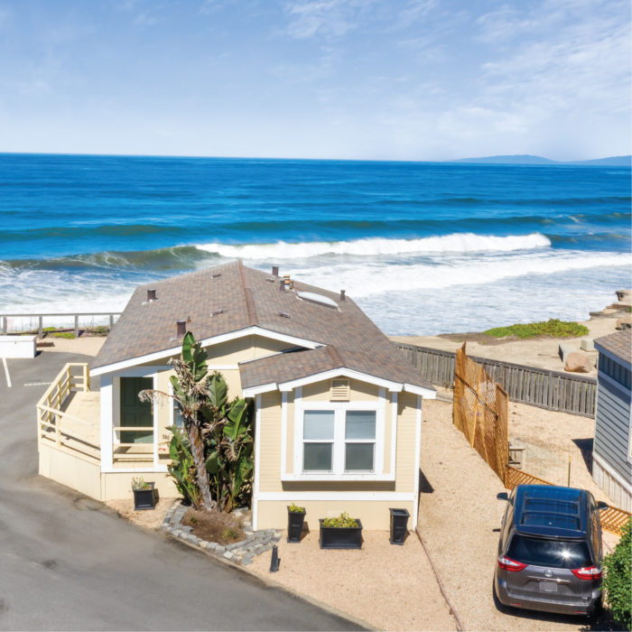 Top view of a beachfront manufactured home with a front lawn, outdoor car space, and the Pacifica, California ocean in the background.