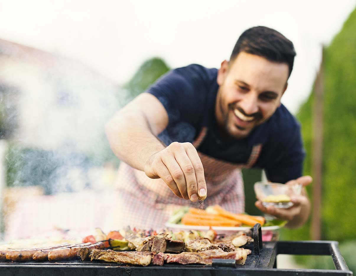 A man grilling in the outdoors, seasons the meat on the grill.